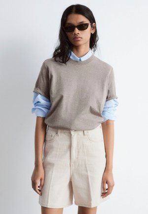 Brown short-sleeve t-shirt layered over a light blue collared shirt, paired with beige high-waisted shorts. Worn with dark sunglasses.