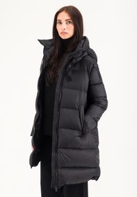 Manteau long noir en tissu brillant et bouffant. Il présente un col haut, une capuche et une fermeture éclair avec des accents élastiques aux poignets et sur les côtés.