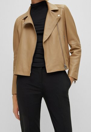 Kobieta w brązowej, skórzanej kurtce motocyklowej na czarnym golfie i czarnych spodniach, stoi z jedną ręką w kieszeni.