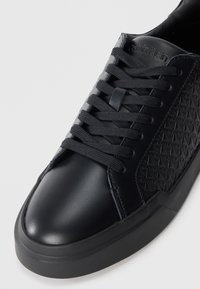 Black leather sneaker featuring a textured side panel with geometric patterns, round toe, and matching black laces on a rubber sole.