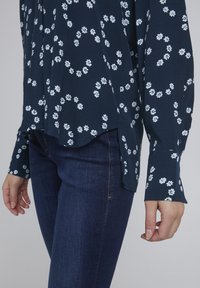 Marineblouse met wit bloemenpatroon, lange mouwen en zijsplitten, gecombineerd met donkere jeans. Glad materiaal, losse pasvorm.