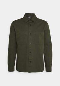 Olijfgroene knoopshirt gemaakt van een gladde stof, met een puntige kraag, twee borstzakken en zwarte knopen.