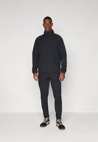 Muž stojaci v čiernom teplákovom súprave s na zips so svetlým pozadím, obutý v čiernych a béžových teniskách na vysokom podpätku.