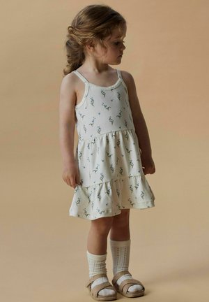Robe blanche imprimée de fleurs avec de fines bretelles et un ourlet à volants en cascade. Associée à des chaussettes blanches jusqu'aux genoux et des sandales beiges.