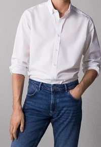 Man wearing a white button-up shirt with rolled sleeves tucked into blue jeans, one hand in pocket against a plain background.