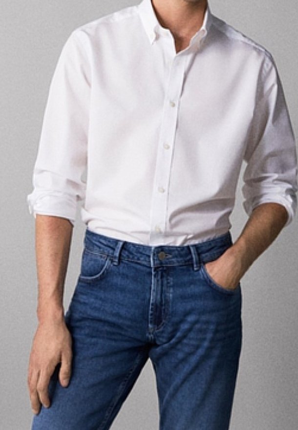 Man wearing a white button-up shirt with rolled sleeves tucked into blue jeans, one hand in pocket against a plain background.