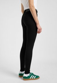 Črne skinny kavbojke z gladko teksturo in visokim pasom, v kombinaciji z zelenimi in belimi supergami z retro dizajnom.