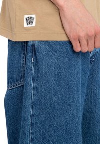 Tan cotton t-shirt with a sewn label featuring black graphics, paired with blue denim shorts displaying a textured surface and faded details.