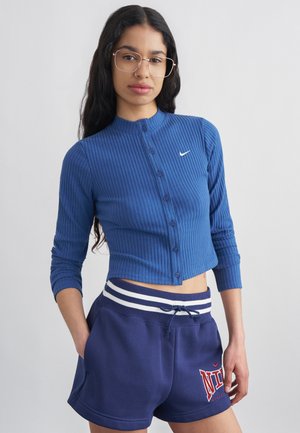 Jeune femme aux longs cheveux foncés, portant des lunettes, un haut bleu côtelé boutonné et un short Nike bleu marine avec des rayures blanches.