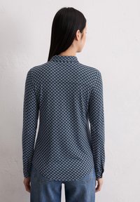 Langarmhemd aus marineblauem Stoff mit einem geometrischen Muster in Hellblau, mit abgerundetem Saum und Kragen. Kombiniert mit blauen Jeans.