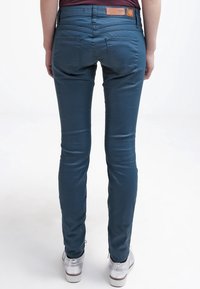 Blauwe slim-fit jeans met een gladde textuur, achterzakken en een leren merkpatch in bruin op de tailleband. Vouwdetails bij de manchetten zichtbaar.