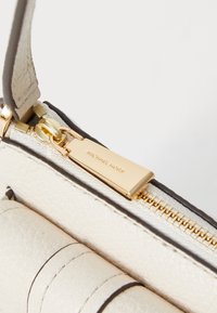 Cream leather handbag featuring textured finish, dark brown accents, a gold zipper, and a metallic brand tag. Simple and modern design elements.