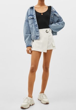 Kvinde iført en cropped denimjakke, sort top, hvid omslagsnederdel med ringdetalje og hvide sneakers stående foran en ensfarvet baggrund.