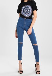 Modré vysoké skinny džíny s roztrhanými koleny, doplněné černým tričkem s grafikou, které má barevný kruhový design. Černé sandály na vysokém podpatku.