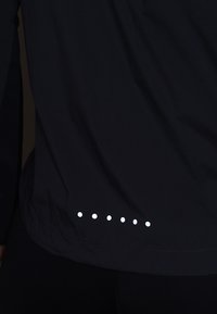 Tissu noir avec une rangée de six petits points réfléchissants blancs près du bord inférieur, porté sur un pantalon noir.
