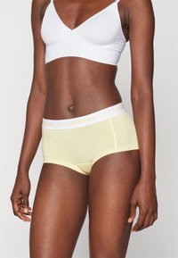 Sous-vêtements hipster en coton jaune avec une taille blanche portant l'inscription "PIECES" en texte jaune clair. Texture douce et design ajusté.