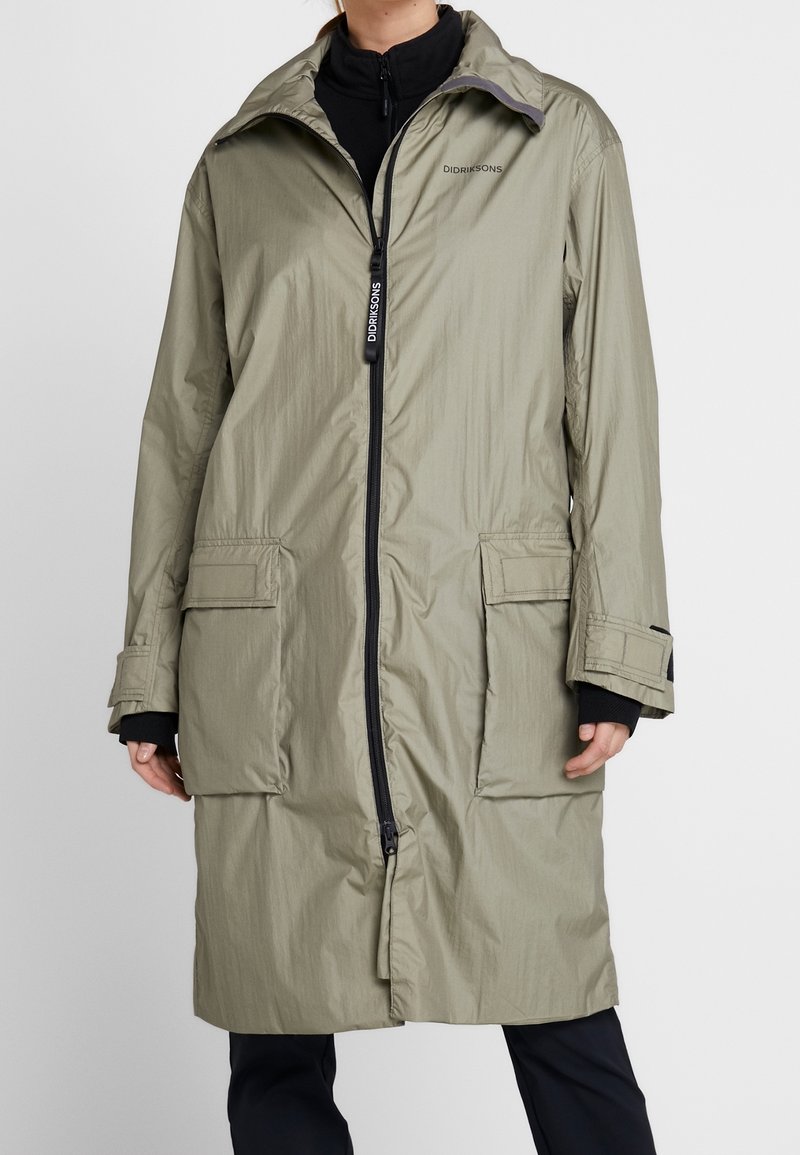 Long manteau vert olive clair avec fermeture éclair à l'avant, poches à rabat et sangles de manche réglables porté sur des vêtements noirs.