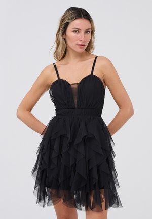 Abito nero con gonna a strati in tulle, corpino aderente, pannello in rete trasparente sul petto e spalline sottili. La texture arricciata crea volume e movimento.