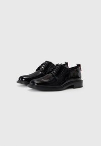 Chaussures en cuir laqué noir avec un design à lacets, bout rond et accents texturés. Elles présentent une languette rouge contrastante au niveau du talon.