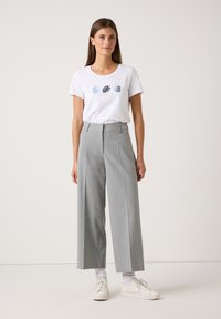 Frau, die auf einem schlichten Hintergrund steht, trägt ein weißes T-Shirt mit blauen Blätterdruck, hellgraue weite Hosen, weiße Socken und weiße Turnschuhe.