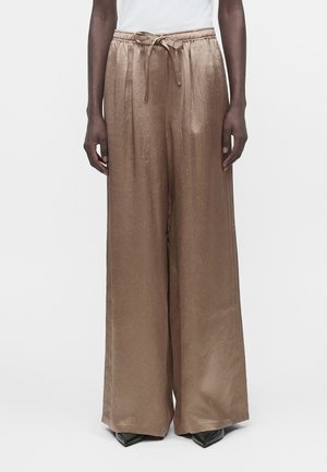 Osoba v širokých, svetlohnedých saténových nohaviciach so sťahovacou šnúrkou v páse a špicatých čiernych topánkach stojí na jednoduchom bielom pozadí.