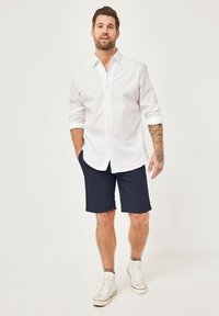 Weißes Langarm-Hemd mit Knöpfen, marineblaue Shorts und weiße High-Top-Sneaker. Kurze, leicht zerzauste Haare und sichtbare Tätowierungen am Arm.