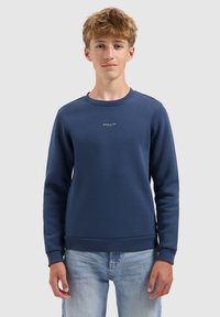 Jongere in een marineblauwe sweater met de tekst "BALLIN'" en lichtblauwe spijkerbroek, staand tegen een effen lichtgrijze achtergrond.