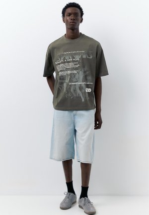 Jeune homme portant un t-shirt graphique Star Wars olive, un short en jean clair, des chaussettes noires et des baskets grises, debout devant un fond blanc.