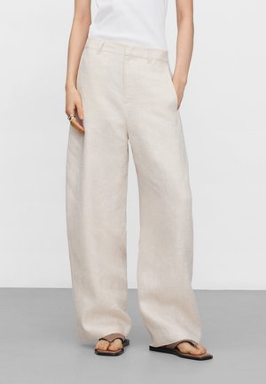Person trägt weite, lockere beige Leinenhose, weißes Oberteil, braune Flip-Flops und Ringe an der linken Hand, posiert mit den Händen in den Taschen.
