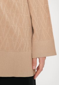 Beige kötött pulóver, texturált gyémántmintával, bordázott mandzsettákkal ésszéllel, sima felülettel és laza illeszkedéssel.