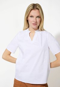Blouse blanche à manches courtes avec un col montant plissé et une ouverture en V. Texture lisse et coupe décontractée, associée à un pantalon marron.