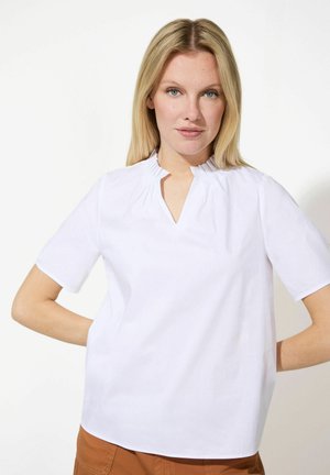 Weiße kurzärmelige Bluse mit einem gefalteten Stehkragen und einem V-Ausschnitt. Glatte Textur und entspannte Passform, kombiniert mit braunen Hosen.