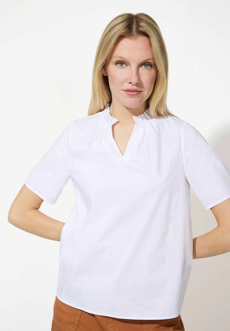 Blouse blanche à manches courtes avec un col montant plissé et une ouverture en V. Texture lisse et coupe décontractée, associée à un pantalon marron.