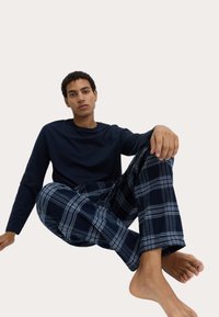 Granatowa koszula z długim rękawem w połączeniu z niebieskimi, kraciastymi spodniami do domu, z białymi paskami. Modelka siedzi na podłodze, boso.