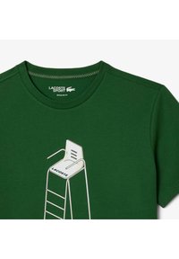 Vihreä puuvillapaita, jossa on valkoinen graafinen kuva pelastustuolista, jossa lukee "LACOSTE" istuinosassa ja yksinkertainen tikasdesign. Normaalimallinen.