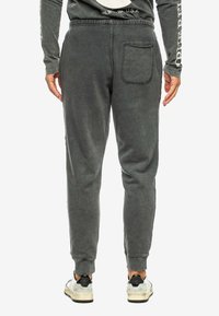Donkergrijze trainingsbroek gemaakt van zacht materiaal, met een achterzak, een elastische tailleband en een taps toelopend ontwerp. Gepaard met witte sneakers.