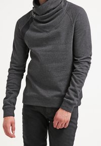 Homme portant un sweat-shirt gris foncé à manches longues avec col asymétrique à fermeture éclair et pantalon noir, debout devant un fond blanc.
