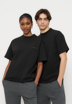 Δύο άτομα φορούν μαύρα T-shirt και γκρι παντελόνια, στέκονται κοντά το ένα στο άλλο, κοιτάζοντας μπροστά με τα χέρια στις τσέπες, μπροστά σε μια απλή, μονόχρωμη επιφάνεια.