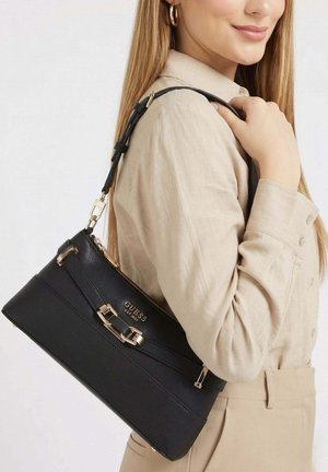 Femme portant une tenue beige tient un sac à main noir Guess avec des accents dorés sur l'épaule, sur fond blanc.