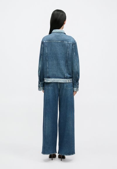 Casaco de ganga com gola azul clara, punhos com botões e costura no peitoral traseiro. Calças de ganga azul escuro de perna larga com bainha limpa.