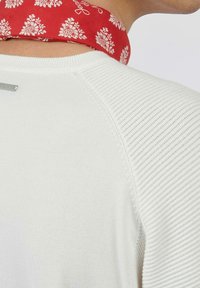 Pull en maille blanche avec des détails côtelés et une écharpe à motifs rouges au niveau de l'encolure, présentant des motifs floraux et une étiquette en métal de marque.