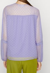 Purple sheer blouse with polka dots, long sleeves featuring lace accents, button closure at neck, and a flowy fit, paired with green pants.