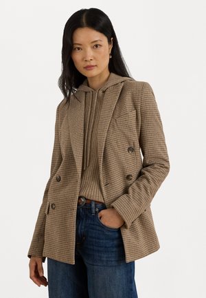 Blazer doppiopetto marrone a motivo houndstooth con rever a punta, tasche frontali e fodera testurizzata indossato sopra un hoodie beige e jeans blu.