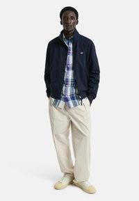 Giubbotto bomber blu navy, camicia a quadri con bottoni in blu, verde e bianco, pantaloni color crema e sneakers bianche con suole beige.