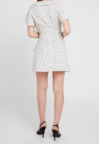 Valkoinen lyhyt mekko mustilla pilkuilla, lyhyet hihat, istuva vyötärö, levenevä hame ja kaulus; yhdistetty mustiin korkeakorkoisiin nilkkureihin.