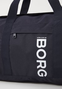 Sötétkék nylon sporttáska vastag pántokkal, elülső zsebbel és feltűnő fehér "BORG" felirattal a könnyű azonosíthatóság érdekében. Texturált anyag.
