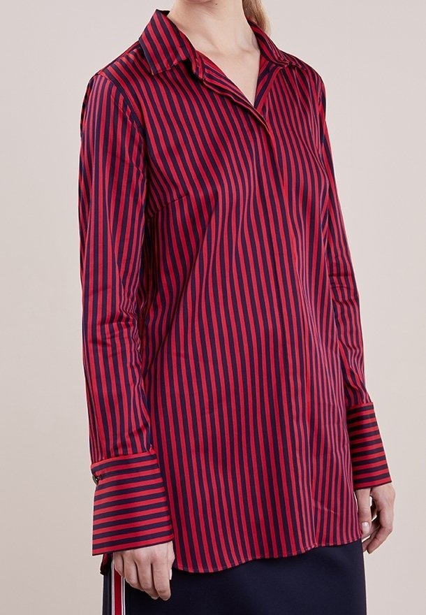 Femme portant un chemisier à rayures verticales rouges et bleu marine avec des manches longues, de larges poignets et un col pointu, debout devant un fond neutre.