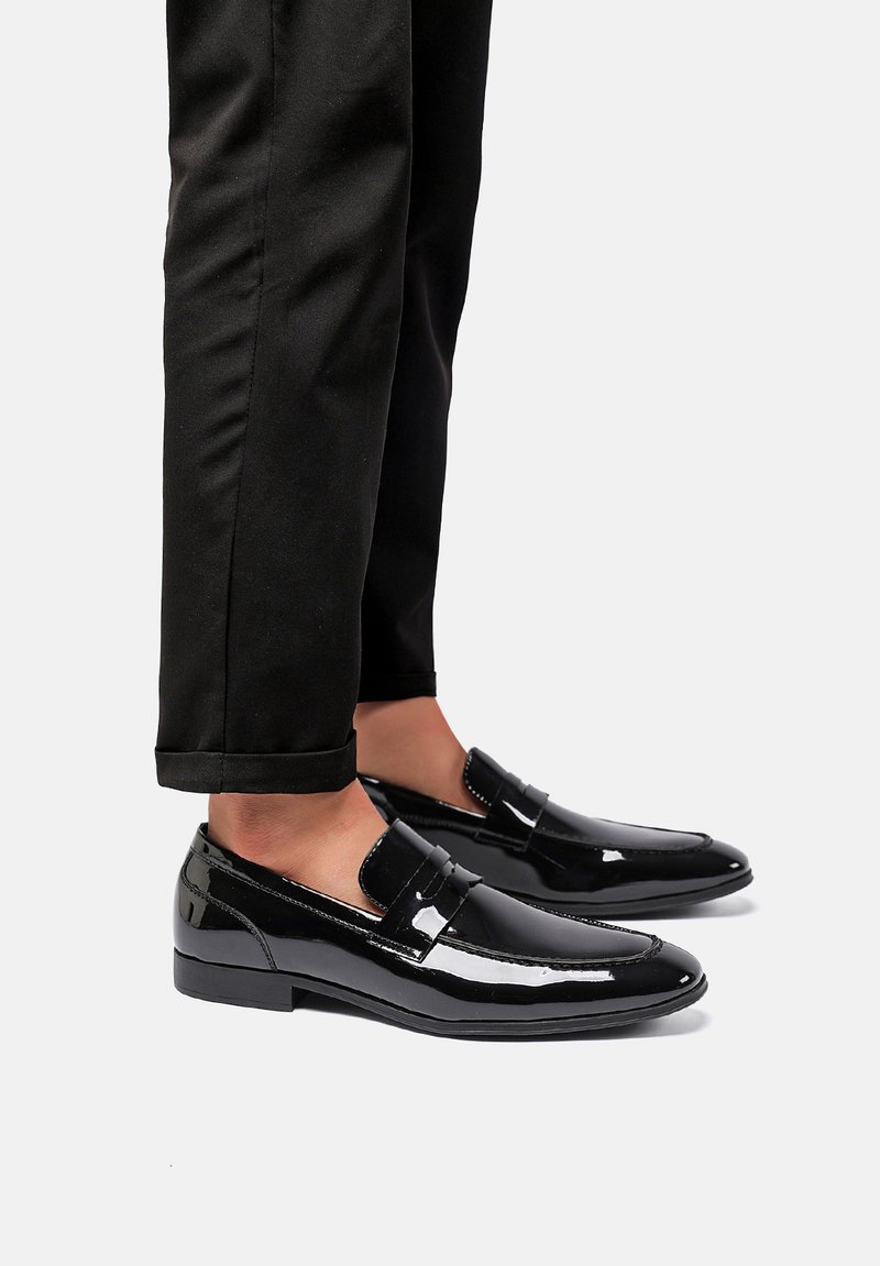 Mocassini neri in vernice con un design elegante, punta arrotondata e un accento decorativo a striscia. Indossati con pantaloni neri con risvolto.