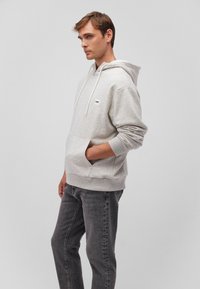 Light grey hooded sweatshirt made of soft fabric, featuring a kangaroo pocket and a small logo tag on the left chest. Paired with dark jeans.