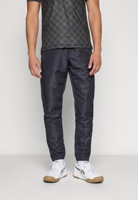 Conjunto deportivo que incluye una camisa de manga corta con un patrón oscuro, pantalones azul marino con puños elásticos y zapatillas deportivas blancas con suela marrón.